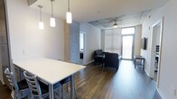 Located at 28 Woolfe St in Charleston's vibrant South of Broad neighborhood, this contemporary single-floor unit showcases a thoughtful blend of modern industrial design and residential comfort. The open-concept layout seamlessly connects the dining and living areas, creating a fluid, spacious feel throughout. Rich, dark hardwood-style flooring runs continuously underfoot, providing a warm contrast to the cool concrete ceiling and accent wall details that give the space a distinctly urban edge. The dining area is anchored by a sleek white table with bar-height seating, illuminated by a trio of modern pendant lights that add both function and style. The living area is defined by large floor-to-ceiling windows fitted with horizontal blinds, inviting an abundance of natural light while offering views to an adjoining balcony. A ceiling fan in the living space ensures year-round comfort. The balcony access through the glazed corner window wall enhances the sense of openness and provides a private outdoor retreat. Neutral white walls throughout keep the space feeling bright and clean, while the interplay of concrete, wood, and metal finishes lends a sophisticated, contemporary character well-suited to the dynamic Charleston lifestyle.