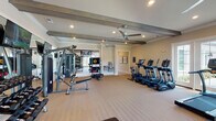 Located at 3215 Midvale Dr in Wilmington, NC, this single-floor fitness center is a comprehensively outfitted facility designed to support a wide range of workout styles and fitness goals. The space features warm neutral walls, light wood-toned flooring, and a ceiling adorned with distinctive decorative wood beams that lend a refined, upscale character. Recessed lighting and multiple ceiling fans ensure the space remains bright and comfortable throughout.

The cardio zone is well-stocked with a row of treadmills, ellipticals, and a stair climber, all positioned near large French doors and windows that invite natural light and offer views to the outdoors. A dedicated strength and functional training area houses a multi-station cable machine, a full rack of dumbbells, a functional training rig complete with TRX suspension straps, kettlebells, and resistance tools, as well as an interactive boxing/reaction training station.

Wall-mounted flat-screen televisions are positioned throughout the space for entertainment during workouts, and full-length mirrors amplify the sense of space while aiding in form and technique. A water fountain is conveniently accessible within the facility. The open, thoughtfully organized layout allows for simultaneous use of multiple equipment zones without congestion, making this fitness center a standout amenity.