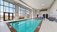 This indoor aquatic facility is designed for both relaxation and recreation, offering a well-appointed environment that balances functionality with comfort. The centerpiece of the space is a generously sized rectangular lap pool with crystal-clear turquoise water, accessible via stainless steel handrails and ladders. Adjacent to the main pool is a separate spa/hot tub, elegantly finished with decorative blue mosaic tile detailing that complements the overall aesthetic. A defining architectural feature of the facility is the expansive floor-to-ceiling window wall along one side, flooding the interior with natural light and offering open views of the surrounding outdoor area. Glass doors provide direct access to an outdoor terrace. The high ceilings, accentuated by exposed HVAC ductwork, lend an industrial yet airy quality to the space. The pool deck is finished with large-format light-toned tiles and is furnished with wicker chaise lounges and modular blue-cushioned seating, creating inviting poolside relaxation areas. A decorative mosaic tile border runs along the lower portion of the walls, adding a vibrant accent throughout the room. The facility also includes changing rooms accessible from the pool deck, making it a fully self-contained aquatic amenity.