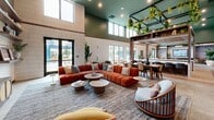 Located at 100 Callan Ave in San Leandro, California, this thoughtfully designed single-floor amenity space offers a welcoming and visually dynamic environment ideal for gathering and relaxation. The interior is defined by dramatically high ceilings painted in a rich sage green, from which an overhead metal grid structure is adorned with cascading live plants, creating a striking botanical installation that anchors the open-plan layout.

The space is divided into several distinct functional zones. A central wet bar features a long stone-topped counter with stylish upholstered barstools, open wood shelving, and a mounted display screen, serving as a natural hub for social interaction. Adjacent to the bar, a formal dining area provides a wooden table with woven cane-back chairs, while a lounge zone offers generous, curved sectional seating in warm terracotta tones paired with low round coffee tables.

A more intimate media nook is outfitted with custom built-in wood shelving and a wall-mounted television, accompanied by sculptural seating in rich leather and velvet. Throughout the space, large floor-to-ceiling windows and glass doors invite abundant natural light and views of the outdoor pool area. Terrazzo and terracotta tile flooring add warmth underfoot, while large-scale artistic murals featuring floral and abstract motifs bring bold character to the walls.