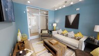 This single-floor space is designed with a contemporary aesthetic, centered around a welcoming living area painted in a bold, calming shade of blue. The room is furnished with a tufted light grey sofa accented by patterned and yellow throw pillows, complemented by a rustic wood coffee table that adds warmth and contrast to the modern surroundings. A sleek leather chair and a side table with a yellow lamp round out the seating arrangement, creating a cohesive and inviting atmosphere.

Track lighting runs along the ceiling, providing adjustable, well-distributed illumination throughout the space. A striking black-and-white geometric artwork anchors one wall, adding a bold visual focal point. The light wood flooring and a patterned area rug in grey and yellow tones tie the color palette together seamlessly.

A notable architectural feature is the floor-to-ceiling glass partition wall with a full-glass door, which separates the main living area from an adjacent room. This transparent divider allows light to flow freely between spaces while maintaining a sense of openness. The overall ambiance is modern, polished, and thoughtfully curated.