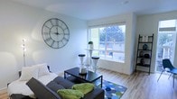 This single-floor space features a well-appointed living area designed with a clean, modern aesthetic. The room is anchored by light hardwood flooring that complements the crisp white walls, creating an airy and open atmosphere throughout. A large window serves as a focal point, flooding the space with natural light while offering views of the surrounding neighborhood and greenery outside.

The living area is thoughtfully arranged with a charcoal sectional sofa accented by lime green throw pillows, adding a pop of color against the neutral backdrop. A sleek black coffee table sits at the center, styled with decorative accessories that enhance the contemporary feel. A geometric-patterned area rug in shades of blue and green defines the seating zone while adding visual interest to the floor.

A standout decorative element is the large wrought-iron Roman numeral wall clock, which adds an industrial-chic touch to the otherwise minimalist space. Near the window, a tall bistro-style table paired with modern stools creates a casual secondary seating or display area. Overall, the space exudes a bright, welcoming ambiance with a balanced mix of modern furnishings and thoughtful design details.