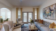 This single-floor residence offers a thoughtfully designed layout with warm wood-look flooring and crown molding throughout. The living room serves as a welcoming centerpiece, featuring French doors flanked by floor-to-ceiling gold drapery that invite natural light and provide outdoor access. A tufted sofa, layered area rug, and bold abstract artwork complete the sophisticated atmosphere.

The dining area occupies a sun-drenched bay window nook, where multiple windows flood the space with natural light. A glass-top round table with a gold base and plush green velvet chairs create an elegant yet inviting setting.

The kitchen is outfitted with gray shaker-style cabinetry, white quartz countertops, a subway tile backsplash, and stainless steel appliances including a French door refrigerator, electric range, and over-the-range microwave. A window above the sink adds additional natural light.

The bedroom features hardwood-style flooring, a generous walk-in closet, and a window dressed with deep navy curtains. The bathroom includes a soaking tub and shower combination with tile surround, a built-in niche, and a white vanity with ample storage.