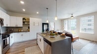 This single-floor residence showcases a contemporary open-concept layout connecting the kitchen, dining, and living areas. The kitchen is outfitted with stainless steel appliances, sleek white cabinetry, a tile backsplash, and a central island with a gray countertop, pendant lighting overhead, and an integrated sink and dishwasher. Wood-look flooring runs throughout the main living areas, adding warmth to the modern aesthetic. The living room is bright and inviting, benefiting from large sliding glass doors and an adjacent window that frame urban skyline views and lead to a private balcony. A ceiling fan enhances comfort in this well-lit space. Multiple bedrooms offer a calm retreat, featuring plush carpeting, natural light from windows with blinds, and neutral tones throughout. The bathrooms are finished with white cabinetry, dark countertops, large mirrors, and wood-look flooring. One bathroom includes a soaking tub and shower combination, while another features a walk-in glass shower. A smart home panel is visible near the kitchen entry, reflecting the unit's modern technological integration. Overall, the space presents a clean, functional, and well-appointed design.