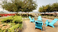 Located at 4040 26th Ave SW in Seattle, WA, this rooftop terrace offers a well-appointed outdoor retreat with a thoughtfully designed layout. A large, striking red shade canopy anchors the main seating area, providing comfortable shelter over multiple bistro-style tables and wicker chairs arranged for casual dining or socializing. Beyond the covered zone, the terrace opens to a spacious sun-drenched deck surfaced with large-format tile pavers. A dedicated outdoor kitchen station, equipped with stainless steel grills and a beverage refrigerator with granite countertops, makes the space ideal for outdoor entertaining. Vibrant teal Adirondack chairs are positioned throughout a beautifully landscaped garden area featuring mature trees, ornamental grasses, flowering shrubs, and raised planter beds that add lush greenery and visual interest. Expansive views of the surrounding Seattle skyline and neighboring structures extend from the terrace, enhancing the open-air ambiance. Warm wood-framed glass doors seamlessly connect the rooftop to an interior lounge space, ensuring easy indoor-outdoor flow. The combination of shaded seating, landscaped gardens, and an equipped outdoor kitchen makes this rooftop terrace a versatile and inviting amenity space.