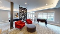 Somerville Parc Lounge is a thoughtfully designed single-floor community amenity space located in Somerville, New Jersey. The lounge offers a welcoming and stylish environment that blends recreational and social functions seamlessly.

The main gathering area features patterned carpeting with a starburst motif and recessed cove lighting that adds a warm, ambient glow to the space. A decorative laser-cut room divider serves as a striking architectural focal point, visually separating lounge zones while maintaining an open feel. Vibrant red accent chairs and a cushioned ottoman provide comfortable casual seating, while additional dining-height tables and chairs offer flexible arrangements for socializing or casual meals. A wall-mounted flat-screen TV anchors one side of the room, and French doors along the perimeter allow natural light to fill the space and provide direct access to the outdoors.

Recreational amenities include an air hockey table and a foosball table, making the lounge an ideal venue for social gatherings and events.

The kitchenette area is finished to a high standard, featuring dark wood cabinetry, white subway tile backsplash, light quartz countertops, a brass-toned undermount sink, and a full-size stainless steel French door refrigerator. The space is both functional and aesthetically refined, complementing the overall modern design of the lounge.