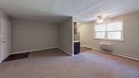 This single-floor unit offers a practical and well-appointed layout designed for comfortable everyday living. The kitchen is a standout feature, showcasing dark espresso cabinetry paired with white subway tile backsplash, light countertops, and a full suite of stainless steel appliances including a gas range, built-in microwave, and refrigerator. Wood-look flooring grounds the kitchen space, adding warmth and durability. The open living area flows naturally from the kitchen and is finished with wall-to-wall carpeting in a neutral tone, creating a cozy and inviting atmosphere. A ceiling fan and a window allowing natural light enhance the comfort of the living space. The bedroom is similarly carpeted and features a window with views of surrounding greenery, baseboard heating, and a wall-mounted HVAC unit for year-round climate control. The bathroom is finished with large-format gray tile on the walls surrounding a full soaking tub with an overhead shower, complemented by white floor tiles and clean, modern fixtures with brushed nickel hardware. Throughout the unit, neutral gray walls provide a versatile backdrop, and consistent baseboard trim ties the aesthetic together cohesively across all rooms.