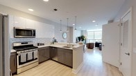 This thoughtfully designed single-floor residence showcases a clean, contemporary aesthetic throughout. The open-concept layout seamlessly connects the kitchen, dining, and living areas, creating a fluid and functional living environment finished with light hardwood-style flooring and neutral warm tones on the walls.

The kitchen is a standout feature, outfitted with white high-gloss upper cabinetry, contrasting dark lower cabinets, stainless steel appliances including a French door refrigerator, microwave, and range, and a striking hexagonal tile backsplash. A peninsula with a white countertop and pendant lighting above provides additional seating and workspace, bridging the kitchen to the dining area.

The dining space comfortably accommodates a full table and chairs, while the adjacent living room opens to a private balcony through floor-to-ceiling sliding glass doors, framing breathtaking panoramic water and hillside views that fill the space with natural light.

The bedroom continues the serene aesthetic, featuring large corner windows that flood the room with daylight and offer sweeping outdoor views. Herringbone-patterned carpet adds texture underfoot, complementing the room's calm, restful atmosphere. The overall design balances modern finishes with a welcoming warmth throughout.