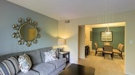 This single-floor residence offers a thoughtfully designed layout that balances comfort and functionality. The living area serves as the heart of the space, featuring plush carpeted flooring and soft, neutral tones complemented by distinctive sage green accent walls that lend a calm, inviting atmosphere. A decorative sunburst mirror adds a stylish focal point, while warm ambient lighting from a table lamp creates a cozy and relaxed environment. The living space flows seamlessly into the dining area through an open passageway, making the layout feel connected and spacious. The dining area continues the sage green color palette, maintaining a cohesive aesthetic throughout the home. It is furnished to accommodate comfortable group dining and is well-lit, adding to the welcoming character of the space. Neutral beige carpeting runs throughout both areas, tying the rooms together with a warm, unified feel. Ceiling vents are integrated discreetly into the design, ensuring climate comfort without disrupting the visual flow. The overall ambiance of this residence is warm, relaxed, and tastefully appointed, making it a comfortable and practical living environment suited for everyday life.