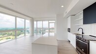 This single-floor residence showcases a cohesive contemporary design, characterized by a crisp all-white palette, light wood-tone flooring, and matte black accents throughout. The open-concept living and kitchen area is the heart of the space, anchored by a generous waterfall-edge island and framed by expansive floor-to-ceiling windows that flood the interior with natural light and offer sweeping elevated city views. The kitchen is thoughtfully appointed with flat-front white cabinetry extending to the ceiling, a striking herringbone tile backsplash, a bold matte black range hood, stainless appliances including a gas range and French door refrigerator, and a built-in microwave. A hallway lined with integrated open shelving connects the living area to the private quarters, maintaining the space's clean aesthetic. The primary bathroom features a floating dual-sink vanity with quartz countertops, matte black fixtures, and a large LED-backlit mirror. A secondary bathroom mirrors this refined style with a single-sink floating vanity and matching finishes. Closet space is provided through a walk-in configuration with wire shelving. Recessed lighting throughout ensures consistent illumination across all areas.