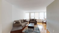 This single-floor residence is thoughtfully laid out to maximize functionality and comfort throughout. The living area serves as the heart of the unit, featuring warm parquet hardwood flooring that extends throughout the space. Large windows fitted with roller shades line the exterior wall, allowing generous natural light while offering privacy control. The seating area accommodates a full sofa and loveseat arrangement centered around a wood coffee table, creating a welcoming space for relaxation.

Adjacent to the living area, the dining space features a round wooden table with upholstered chairs beneath a statement drum pendant light, with a window providing additional natural light. The kitchen is efficiently designed with teal-toned lower cabinetry, dark granite countertops, white glass-front upper cabinets, a subway tile backsplash, and stainless steel appliances including a gas range and microwave.

The bedroom offers a comfortable, quiet retreat with parquet flooring, neutral walls, and a large window with outdoor views and roller shades. The bathroom is clean and functional, featuring a full soaking tub with a tiled surround, a curved dark vanity with a white basin, and a mirrored medicine cabinet.