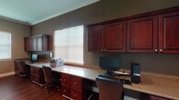 This single-floor office space is thoughtfully designed to support a productive work environment, combining functional built-ins with refined finishes. The room is anchored by an expansive L-shaped built-in desk with a granite-style countertop, offering ample workspace for multiple users simultaneously. Rich, dark cherry wood cabinetry lines the walls both above and below the desk surface, providing generous storage through a combination of upper wall-mounted cabinets and lower drawer units with brushed nickel hardware. The warm-toned hardwood flooring runs throughout, adding a polished, professional character to the space. Neutral taupe-gray walls serve as a clean backdrop, while crown molding along the ceiling lends an architectural refinement. Natural light filters in through multiple windows dressed with horizontal blinds, keeping the space bright yet glare-controlled. Recessed lighting in the ceiling supplements the natural light, ensuring well-distributed illumination across the workspace. A set of framed wall art pieces adds a decorative touch without detracting from the room's functional focus. The space also includes a closet, offering additional storage. Overall, this office presents a cohesive, well-organized environment suited for both focused individual work and collaborative use.
