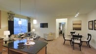 Located at 1982 Route 70 East in Cherry Hill, New Jersey, this single-floor residence offers a thoughtfully designed open-concept layout that maximizes both space and functionality. The interior is anchored by a fluid connection between the kitchen, living, and dining areas, creating an ideal environment for both everyday living and entertaining. The kitchen features dark granite countertops that provide a bold contrast to the light-toned walls and neutral carpeting found throughout the space. A stylish pendant light hangs above the kitchen island, adding a modern accent. The living area is generously proportioned, accommodating a comfortable seating arrangement centered around a wall-mounted television, and benefits from abundant natural light streaming in through large windows dressed with floor-length curtains. A cozy accent chair complements the main seating, while a compact dining set with sleek dark chairs sits nearby, ideal for casual meals. A hallway visible from the main living area leads to additional rooms, suggesting a well-organized floor plan. The overall ambiance is warm and inviting, with a soft, neutral color palette, smooth transitions between spaces, and consistent finishes that lend the interior a cohesive and polished character throughout.