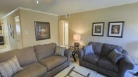 This single-floor residence offers a cohesive and comfortable living environment designed with both style and functionality in mind. The living room serves as the heart of the space, featuring warm neutral wall tones that create a welcoming ambiance. Two plush sofas in complementary shades of gray and taupe are arranged to encourage conversation, each accented with decorative throw pillows. A glass-topped accent table sits between the seating, topped with an elegantly styled table lamp that casts a soft, warm glow throughout the room. Crown molding along the ceiling adds a refined architectural touch, while framed wall art featuring classic still-life and botanical motifs lends a timeless character to the space. A light-toned area rug anchors the seating arrangement, and a doorway visible from the living area provides a glimpse into the adjoining spaces, suggesting a well-connected and functional floor plan. The overall ambiance is calm and inviting, with thoughtful attention to detail evident in the décor choices. Natural and artificial lighting work together to keep the space feeling bright and livable, making it a comfortable and practical single-floor home.