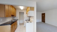 Sharon Arms is a single-floor residence that combines practical design with comfortable living. The kitchen is a standout feature, equipped with warm honey-toned wooden cabinetry that provides ample storage space. White appliances, including a coil-top electric range, overhead microwave, refrigerator, and dishwasher, are neatly arranged to create an efficient workflow. A dark countertop with an integrated sink adds contrast to the cabinetry, while light tile flooring keeps the space bright and easy to maintain. The kitchen opens seamlessly into the adjacent living area, which features neutral carpet flooring, creating a cohesive flow between spaces. The open-concept connection between the kitchen and living area allows for easy interaction and a sense of openness throughout the main living spaces. Neutral wall tones throughout contribute to a clean, understated aesthetic that allows for easy personalization. A doorway visible from the kitchen suggests additional rooms beyond, offering private retreats within the home. Overall, Sharon Arms presents a straightforward and well-organized layout suited for comfortable, everyday living.