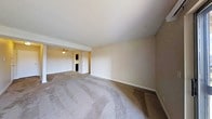 This single-floor space presents an open and functional layout defined by neutral beige walls and wall-to-wall carpeting that adds a sense of warmth underfoot. The main living area is generously proportioned, offering ample room for a variety of furniture arrangements. A textured white ceiling adds subtle visual interest overhead, while clean white baseboards provide a crisp contrast against the neutral walls.

An open passage connects the main living area to a secondary space, which features a ceiling fan with a light fixture, providing both ventilation and overhead lighting. Beyond this area, a glimpse of what appears to be a kitchen or utility space is visible, suggesting a practical and connected floor plan.

The layout flows naturally from one area to the next, creating a cohesive and easy-to-navigate living environment. The overall ambiance is calm and understated, with a blank-canvas quality that allows for personalization. The absence of obstructions between rooms enhances the sense of openness throughout the space, making it feel inviting and adaptable to a range of uses.