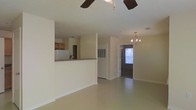 Located at 616 Highway 90 in Waveland, Mississippi, this single-story property offers a practical and open floor plan designed for comfortable living. The main living area is generously sized, featuring smooth, light-toned flooring and neutral wall colors that create a bright and airy atmosphere. A ceiling fan overhead provides efficient air circulation throughout the space.

The kitchen is accessible via a half-wall pass-through counter finished with a stone-style surface, offering both separation and visual connectivity to the living area. Wood-toned cabinetry and a white refrigerator are visible, giving the kitchen a classic, functional character.

Toward the rear of the living space, an elegant chandelier adds a touch of refinement, illuminating the transition into what appears to be a hallway or additional room. Natural light filters in through a window at the far end, enhancing the open feel of the interior.

The layout flows smoothly from one area to the next, with clean sight lines running the length of the home. White doors and trim provide crisp contrast against the warm neutral walls, contributing to a cohesive and well-maintained aesthetic throughout this accessible, single-level property.