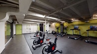 Located at 512 W Ormsby Ave in Louisville, Kentucky, this fitness center offers a well-equipped, single-floor workout facility designed to support a variety of exercise routines. The space features an open, industrial aesthetic with exposed structural beams and ductwork overhead, complemented by bright fluorescent lighting that evenly illuminates the entire floor. Durable rubber flooring covers the workout areas, providing a safe and functional surface for both cardio and strength training activities. The facility is outfitted with a comprehensive selection of equipment, including spin bikes, treadmills, elliptical machines, and a multi-function cable strength machine, as well as a flat bench and an assortment of exercise balls and resistance equipment along the walls. Bold yellow-green accent doors provide pops of color against the neutral concrete block walls, lending the space a modern, energetic character. A hand sanitizer station and informational bulletin board are conveniently positioned near the entrance. The layout allows for ample circulation space between equipment, supporting a comfortable and organized workout environment. An adjacent glass-partitioned area is visible from the main floor, suggesting additional amenity space nearby. The overall design prioritizes functionality and accessibility for users of all fitness levels.