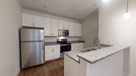 This single-floor unit showcases a clean, contemporary design throughout its thoughtfully arranged layout. The kitchen is a standout feature, offering crisp white shaker-style cabinetry, granite countertops, and a full suite of stainless steel appliances including a refrigerator, range, microwave, and dishwasher. A peninsula with a built-in sink provides additional prep space and casual seating potential, while pendant lighting adds a modern finishing touch. Rich hardwood-style flooring runs throughout the main living and kitchen areas, creating a warm and cohesive aesthetic.

The bathroom continues the refined design language with granite-topped vanity cabinetry, hardwood-style flooring, and a clean, neutral color palette that lends a bright and airy feel. The bedroom is carpeted for comfort and features a large window with blinds that invites natural light and offers views of surrounding greenery, creating a peaceful retreat.

The overall ambiance of the space is modern and well-appointed, with consistent finishes across rooms that contribute to a polished, move-in-ready feel. Neutral walls and carefully selected materials throughout ensure a versatile backdrop suitable for a variety of interior styles.