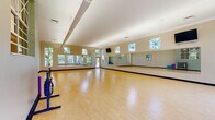 This single-floor fitness and dance studio is designed to accommodate a wide range of movement-based activities, from group exercise classes to dance instruction. The main studio space is expansive and open, featuring smooth, light-toned hardwood flooring that provides an ideal surface for both high-impact workouts and dance practice. Soaring ceilings with exposed ductwork lend an airy, industrial-inspired aesthetic, while full-length wall mirrors along multiple walls allow participants to monitor their form and enhance the sense of space. Natural light floods the studio through a generous arrangement of large windows and decorative multi-paned windows positioned at upper wall levels, offering views of lush greenery outside and creating a bright, energizing atmosphere. Glass-paneled entry doors further connect the interior to the outdoors. Wall-mounted flat-screen televisions are positioned for clear sightlines across the studio floor, supporting instructor-led programming. A secondary, more intimate room off the main studio features the same warm hardwood flooring, a neutral color palette, and a single framed window, making it well-suited for smaller group sessions, yoga, or mat-based fitness. Throughout the facility, navy blue baseboards provide a clean, defined contrast to the warm floor tones and light-colored walls, contributing to a polished, professional environment purpose-built for fitness and wellness.