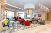 Located on Schering Drive in Union, NJ, this contemporary amenity space delivers a sophisticated and dynamic environment designed for both relaxation and entertainment. The grand lobby lounge immediately impresses with soaring ceilings adorned with sleek linear LED lighting that runs in dramatic parallel lines across a soft teal-toned ceiling. Floor-to-ceiling glass walls and large windows flood the space with abundant natural light while offering open views of the surrounding exterior grounds. The entrance area features a striking sculptural pendant light and vibrant, artistically patterned carpeting in shades of blue, teal, and grey that flows throughout the space, lending it a cohesive, gallery-like aesthetic. Comfortable seating arrangements — including plush sectional sofas, accent chairs in mustard yellow and white, and sleek coffee tables — are thoughtfully positioned throughout the lounge. A wall-mounted flat-screen TV anchors the main seating area, while a staircase and bar-height seating are visible in the background. The entertainment wing transitions into a game room featuring decorative mosaic-tiled columns, pendant globe lighting, a billiards table, and classic stand-up arcade cabinets. A dedicated multi-sport simulator room rounds out the amenities, complete with a large projection screen and artificial turf flooring, supporting golf, football, and more.