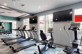 This expansive fitness center is thoughtfully designed to support a comprehensive workout experience across multiple functional zones. The space features a striking herringbone-patterned carpet in tones of teal, gray, and beige that runs throughout, lending a modern, cohesive aesthetic to the entire facility.

The cardio zone is outfitted with a row of TRUE treadmills, elliptical trainers, stationary bikes, and a Freemotion cable machine, all positioned near large windows that offer views of an outdoor pool area with red umbrella-shaded seating. Wall-mounted flat-screen televisions are positioned throughout for entertainment during workouts. Ceiling fans ensure consistent air circulation across the space.

A dedicated strength training area houses a multi-station cable machine, weight benches, and selectorized resistance equipment. Full-length mirrors amplify the sense of space and allow for proper form checks. Accent walls painted in deep teal, paired with abstract artwork, add a bold visual contrast to the otherwise neutral gray palette.

A separate studio zone provides space for cycling classes, featuring smart stationary bikes with integrated screens, floor-length mirrors, and ample open floor space for stretching or mat work. A free weights area includes a color-coded dumbbell rack, medicine balls, stability balls, and kettlebells, rounding out the facility's versatile offerings.