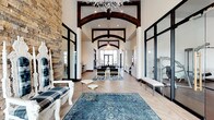 Located at 5252 Riggins Run in Dublin, Ohio, this sophisticated single-floor clubhouse delivers an impressive blend of rustic and contemporary design. The grand entrance immediately captures attention with its soaring vaulted ceiling adorned with dramatic dark wood arched beams and an eye-catching sculptural chandelier that serves as a striking focal point. Elegant herringbone-patterned light wood flooring flows throughout the entire space, grounding the design with warmth and visual continuity.

The main hall opens into a spacious lounge area furnished with leather seating, accent tables, and richly toned area rugs, creating an inviting atmosphere for gathering and socializing. A natural stone accent wall with a fireplace anchors one side of the space, adding texture and a sense of rustic character to the otherwise modern aesthetic.

Floor-to-ceiling black-framed glass walls enclose a fully equipped fitness center, maintaining an open, airy feel while clearly delineating the workout space. The transparency of the glass partitions allows natural light to pass freely throughout the interior, enhancing the sense of openness. Additional amenities include built-in cabinetry and counter space, ideal for entertaining or everyday convenience.

Large black-framed windows and glass entry doors frame views of the outdoors, flooding the interior with natural light. The overall ambiance is refined yet approachable, combining high-end finishes with functional design throughout.