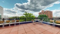 This rooftop terrace offers an open-air retreat elevated above the surrounding urban landscape. The floor is finished with large-format terracotta tiles that extend across the generous, unobstructed deck area, creating a warm and cohesive aesthetic underfoot. A sturdy metal railing system with cable infill runs along the perimeter, ensuring safety while preserving sweeping views of the city skyline, neighboring brick and modern buildings, and a lush canopy of mature trees.

The terrace is furnished with multiple seating arrangements, including slatted wood-and-metal chairs paired with rectangular tables, as well as additional bench-style seating positioned to take advantage of the views. The furnishings reflect a clean, contemporary design consistent with the terrace's modern architectural character.

Access to the terrace is provided through a glass-paneled door framed by panel-clad walls in neutral gray and bold orange accents, lending a contemporary, urban feel to the entry point. Wall-mounted speakers and utility fixtures are also present, suggesting the space is equipped for social gatherings or events. Open to the sky, the terrace receives abundant natural light throughout the day, making it a versatile and inviting outdoor space.