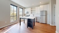 This single-floor space showcases a thoughtfully designed interior with a clean, contemporary aesthetic throughout. The kitchen serves as a central highlight, featuring white shaker-style cabinetry, stainless steel appliances including a range, microwave, and refrigerator, and a classic white subway tile backsplash. A freestanding center island with a crisp white countertop and dark base provides additional prep space and storage. Bold black-framed windows line the kitchen walls, flooding the area with natural light and offering pleasant outdoor views of surrounding greenery. Light-toned hardwood flooring runs throughout the main living areas, contributing to a bright and airy atmosphere.

The bathroom presents a clean, functional design with wood-look flooring, a full tub-and-shower combination finished with white tile surround, and matte black fixtures that add a modern contrast. A white vanity completes the space with understated elegance.

An additional room features plush carpeting, neutral wall tones, and a wall-mounted sconce with a dark bronze finish, creating a warm, intimate ambiance. Recessed lighting throughout the main areas ensures consistent, even illumination. The overall design balances modern finishes with comfortable, livable details.