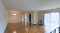 This single-floor residence offers a clean, straightforward layout designed for comfortable everyday living. The main living area is expansive and open, showcasing warm hardwood flooring that extends throughout the space, lending a sense of continuity and warmth. Neutral-toned walls keep the interior bright and versatile, ready to accommodate a variety of décor styles.

An elegant chandelier anchors the dining area, providing a touch of refinement to the otherwise understated aesthetic. Natural light filters in through a sliding glass door dressed with vertical blinds, offering access to the outdoors and brightening the living space. A wall-mounted air conditioning unit ensures year-round comfort, complemented by baseboard heating along the perimeter.

The layout flows naturally from the main living and dining areas toward additional rooms visible through interior doorways, suggesting well-separated functional spaces. A dark-toned refrigerator is visible in what appears to be a transitional or kitchen area, hinting at practical amenities nearby.

Overall, the space presents a functional, well-maintained interior with classic finishes, ample room to move, and a layout that balances openness with defined living zones.