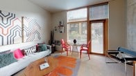 This single-floor unit is thoughtfully organized into distinct living zones. The open-concept kitchen is outfitted with light maple cabinetry, dark countertops, and black appliances including a full-size refrigerator, electric range, and dishwasher. Track lighting illuminates the space, which flows directly into a compact dining area furnished with a glass-top table and contemporary metal stools, all anchored by light hardwood flooring.

The living room benefits from floor-to-ceiling windows with wood-framed surrounds, allowing substantial natural light and views of the surrounding exterior. The generous ceiling height adds to the open, airy feel of this space.

The bedroom features carpeted flooring, tall ceilings, and a built-in closet system with dual sections, providing ample storage. A hallway connecting the bedroom to the bathroom includes additional wire shelving for organizational use.

The bathroom is equipped with a dark-countertop vanity with maple cabinetry, a shower with curtain enclosure, and light hardwood flooring. An in-unit stacked washer and dryer is conveniently located in the connecting hallway. Neutral wall tones throughout create a cohesive, modern aesthetic.