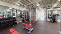 The Merrifield Fitness Center at Dunn Loring Station, located at 3180 Prescott Drive in Vienna, Virginia, is a thoughtfully designed, single-floor fitness facility offering a comprehensive workout environment. Guests enter through a bright, clean foyer finished with light wood-look tile flooring, a branded welcome mat, and a seating bench — setting a welcoming tone before stepping into the main fitness space.

The fitness floor is expansive and open, featuring durable rubber flooring throughout and an exposed structural ceiling with integrated lighting that lends an industrial-modern aesthetic. Large windows lining the perimeter flood the space with natural light and offer views of the surrounding greenery, creating an energizing atmosphere.

The facility is divided into well-defined workout zones. A strength training area includes squat racks with red benches, a dumbbell rack, cable machines, and a multi-functional cable cross station. A dedicated selectorized machine zone houses a row of modern resistance machines, complemented by large glass panels with motivational graphics. Functional training is supported by stability balls, medicine balls, and open floor space. A cardio section with stationary bikes is visible through the entrance doorway. Full-length mirrors enhance the sense of space throughout. The facility is clean, modern, and comprehensively outfitted for a full-body fitness experience.