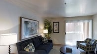 This single-floor space presents a comfortable and thoughtfully arranged living area that balances functionality with a warm, inviting ambiance. The living room serves as the focal point, featuring neutral-toned walls that provide a versatile backdrop for décor. A textured ceiling adds subtle architectural interest overhead, while light-toned flooring anchors the room with a clean, cohesive look.

The space is furnished with a deep navy sofa accented by patterned throw pillows, complemented by a round dark coffee table that adds contrast to the lighter surroundings. A large, framed abstract artwork dominates one wall, contributing a contemporary artistic touch, while a smaller botanical print adds visual balance nearby. A potted palm plant introduces a natural, organic element to the corner of the room, enhancing the overall freshness of the space.

A sliding glass door fitted with vertical blinds allows natural light to filter into the living area while providing direct access to an outdoor space, contributing to an open and airy feel. A floor lamp positioned near the seating area offers warm supplemental lighting, creating a cozy atmosphere during evening hours.