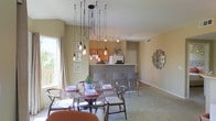 This single-floor space showcases a thoughtfully designed open-concept layout that blends dining and kitchen areas into one cohesive living environment. The dining area serves as an immediate focal point, anchored by a circular glass-top table surrounded by a mix of wooden and upholstered chairs. Above it hangs a dramatic, artistic multi-pendant chandelier with exposed Edison bulbs cascading at varying heights — a bold design statement that adds warmth and character to the space. Natural light pours in through a large window that overlooks an outdoor area, enhancing the airy and welcoming atmosphere. The kitchen is positioned just beyond the dining space, separated by a raised breakfast bar with seating, creating a natural yet open division between the two areas. Warm-toned wood cabinetry and a full-size refrigerator are visible from the dining area, reinforcing the connected feel of the layout. Track lighting above the kitchen ensures functional illumination throughout. Neutral-toned walls and light carpeting provide a clean, versatile backdrop throughout, complemented by tasteful décor accents including framed wall art and a decorative sunburst mirror. The overall ambiance is modern yet inviting, balancing everyday functionality with distinctive design touches.