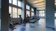This fitness center offers a thoughtfully designed workout environment that combines functionality with an open, airy atmosphere. The space is defined by its soaring ceiling height, which creates a sense of expansiveness throughout the room. A row of large windows lines one wall from floor to near ceiling, flooding the interior with natural light and offering views of surrounding greenery and the outdoors, enhancing the overall workout experience.

The flooring features a durable tile pattern in neutral grey tones accented with blue square insets, providing both practicality and visual interest underfoot. Along the windowed wall, a lineup of cardio equipment—including treadmills, stationary bikes, and rowing or elliptical machines—is neatly arranged, allowing users to exercise while enjoying the natural light and outdoor views.

Toward the rear of the space, additional strength and multi-use equipment is visible, offering a well-rounded selection of workout options. The ceiling incorporates recessed lighting and pendant fixtures, supplementing natural light for consistent illumination throughout all hours. The walls are finished in a contemporary dark grey palette, lending a modern, clean aesthetic to the overall design.