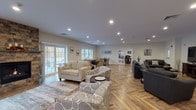Located at 1 Regency Village Way in Merrimac, Massachusetts, this single-floor community clubhouse offers a generously sized, well-appointed gathering space designed for social interaction and relaxation. Throughout the entire space, striking herringbone-patterned hardwood flooring ties each area together, complemented by recessed lighting and neutral, warm-toned walls.

The expansive main lounge features multiple seating arrangements, including plush sofas and accent chairs, with French doors and large windows that flood the room with natural light and provide views of an outdoor deck. Adjacent to the lounge is a dedicated media area furnished with comfortable sectional sofas, a large wall-mounted television, and ample room for group gatherings.

A card and game room offers multiple round tables surrounded by tufted, upholstered chairs, ideal for games and socializing. Large windows in this area frame views of the surrounding wooded landscape. A separate bar area includes a handsome dark wood bar with seating, a wall-mounted TV, and a wet bar with white cabinetry, glass-front storage, and granite countertops. A nearby pub-style room with high-top tables adds a casual entertainment option. The space also includes a well-equipped catering kitchen featuring rich wood cabinetry, granite countertops, and modern appliances. Sliding glass doors in a quiet sitting nook provide direct access to the outdoor deck.
