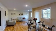 This single-floor space offers a clean, well-appointed layout centered around an open-concept living and dining area. The warm-toned hardwood floors run throughout, lending a cohesive and inviting feel to the entire space. Recessed lighting in the smooth white ceiling provides consistent illumination, complementing the generous natural light that streams in through multiple windows fitted with white shades and classic trim detailing.

The living area is comfortably arranged with a light-colored sofa set accented by decorative throw pillows, and the neutral beige walls are adorned with framed artwork, including prints with a classic Parisian theme, adding a touch of personality to the space.

Seamlessly connected, the dining area features a glass-top table with elegant wrought-iron chairs, set with formal place settings and accented by a vibrant floral centerpiece. The combination of the glass table and the open floor plan contributes to a sense of spaciousness and airiness throughout.

Overall, the space presents a harmonious blend of functional design and comfortable living, with natural light, warm flooring, and thoughtful décor creating an inviting and well-balanced environment suitable for both everyday living and entertaining.