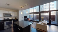 This contemporary two-floor residence showcases a thoughtfully designed open-concept layout with a seamless flow between the kitchen and living areas. The main level is anchored by a dramatic wall of floor-to-ceiling windows that flood the living room with natural light and offer views of the surrounding urban landscape. Soaring ceilings add a sense of grandeur to the space, while dark hardwood flooring runs throughout, providing a striking contrast to the bright white walls.

The well-appointed kitchen features a two-toned design with dark lower cabinetry and white upper cabinets, complemented by dark granite countertops, stainless steel appliances, and a central island with bar seating illuminated by pendant lighting. A half bathroom on the main level adds convenience for guests.

The upper level houses the bedrooms, each offering a tranquil retreat with clean lines and neutral tones. The primary bedroom includes access to a walk-in closet and is served by a full bathroom featuring large-format gray tile flooring, a tub-shower combination with classic white subway tile surround, and dark vanity cabinetry. A second bedroom provides additional flexible living space. The overall ambiance is modern, urban, and refined.