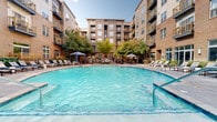 Located at 730 Hawthorne Lane in Charlotte's vibrant Elizabeth neighborhood, this outdoor amenity space centers around a generously sized resort-style swimming pool with clear, blue-tinted water. The pool is framed by a spacious concrete deck outfitted with rows of lounge chairs and chaise lounges, ideal for relaxing in the Carolina sun. Shaded retreats are provided by elegant navy blue canopy cabanas, each furnished with comfortable seating, offering a more private lounging experience. Additional umbrella-shaded seating areas are scattered throughout the deck for added comfort. The courtyard is enclosed on multiple sides by a handsome multi-story building featuring a blend of warm brick and neutral stucco facades, with private balconies featuring gold-toned railings overlooking the amenity area. Mature trees and carefully maintained landscaping add natural beauty and shade to the space, creating a welcoming and verdant atmosphere. Decorative iron fencing defines the perimeter of the courtyard, while pathway lighting ensures the space remains functional and inviting into the evening hours. The overall ambiance is that of a well-appointed, resort-inspired outdoor retreat nestled within an upscale urban residential community in Charlotte, NC.