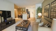 This single-story residence showcases an open-concept layout that connects the living, dining, and kitchen areas into one cohesive, flowing space. The living room is anchored by a plush neutral sofa paired with a rustic wooden coffee table, creating a warm and inviting focal point. Track lighting runs throughout the ceiling, providing consistent, adjustable illumination across all areas. The kitchen features dark cabinetry contrasted against light countertops, with a peninsula that doubles as a casual dining or prep space. Adjacent to the kitchen, a dedicated dining area accommodates a table with seating, framed artwork adorning the warm-toned walls. The flooring transitions between plush carpet in the living areas and hard surface flooring near the kitchen and entry, adding both comfort and practicality. A tall decorative plant near the living area introduces a natural, organic element to the interior. The warm beige and taupe color palette, combined with layered lighting from both track fixtures and a floor lamp, creates an overall ambiance that feels cozy, well-appointed, and thoughtfully arranged throughout.