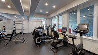 This fitness center offers a comprehensive workout environment designed to accommodate a wide range of exercise routines. The space is thoughtfully organized into distinct zones, each dedicated to a specific type of training. The cardio area lines one side of the room and features multiple treadmills, elliptical machines, and stationary bikes, including recumbent models. Large windows along the walls allow natural light to fill the space, creating an open and energizing atmosphere.

The strength training section is equally well-appointed, housing a multi-station cable machine, a squat rack with weight plates, an adjustable bench press, and a dedicated free weights area. The free weights zone includes a full rack of dumbbells in varying sizes, kettlebells, and a selection of medicine balls, all neatly organized on racks. Large mirrors line this section of the wall, enhancing the sense of space and allowing users to monitor their form.

Underfoot, durable rubber flooring covers the entire area, providing both safety and sound absorption. The ceiling features a distinctive tiered design with recessed lighting that ensures consistent illumination throughout. The overall aesthetic is clean and modern, with a white and grey color palette that contributes to a focused, professional workout environment.