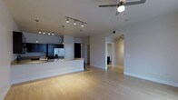 This single-floor space in Dallas, TX, showcases a contemporary open-concept design that integrates the kitchen and living areas into one cohesive, light-filled environment. Light hardwood flooring spans the main living space, contributing to a clean, modern aesthetic throughout.

The kitchen is well-appointed with dark cabinetry, stainless steel appliances including a refrigerator and range, and a granite-topped peninsula that serves as both a prep surface and a casual dining or entertaining area. A pendant light above the peninsula complements the track lighting overhead, providing layered, functional illumination across the kitchen zone.

The open living area benefits from a ceiling fan, enhancing comfort and airflow. Neutral gray walls create a versatile backdrop, while the seamless transition between spaces gives the layout a spacious, unobstructed feel. A hallway visible from the main area leads to additional rooms, suggesting a thoughtful and practical floor plan. The overall ambiance is modern and understated, with clean lines and a neutral palette that offers a welcoming, move-in-ready environment.