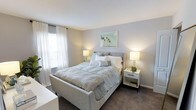 This single-floor residential space offers a thoughtfully designed and inviting interior with a clean, contemporary aesthetic. The bedroom serves as a standout feature, showcasing a calming neutral color palette of soft grays and whites that creates a serene and restful atmosphere. Plush wall-to-wall carpeting adds warmth underfoot, complementing the room's cozy character. A generously sized bed anchors the space, flanked by matching white nightstands fitted with warm-toned table lamps that cast a soft, welcoming glow. Large windows dressed in sheer white curtains allow an abundance of natural light to filter into the room, enhancing its airy and open feel. A framed tropical-themed artwork above the bed adds a subtle decorative touch, introducing a hint of color and personality to the otherwise understated décor. The smooth, light-toned walls and clean ceiling finish contribute to the overall sense of brightness and order throughout the space. The layout is practical and well-proportioned, making efficient use of the available floor area while maintaining a comfortable and uncluttered environment. Overall, the space presents a polished, move-in-ready interior that balances functionality with a relaxed, modern sensibility.