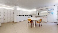 This shared laundry facility offers a clean, bright, and efficiently organized space designed to accommodate high-volume use. The layout is divided into two functional areas, all finished with light-toned tile flooring and a textured ceiling with soft ambient cove lighting that creates an evenly lit, comfortable environment.

The main laundry area is lined with a full row of commercial-grade top-loading washing machines and dryers, each equipped with digital payment and control panels. Above the machines, a continuous run of white upper cabinets provides generous storage space. Flanking the appliances, rows of individual white storage lockers — arranged in a grid pattern across multiple walls — offer secure personal storage for residents.

A centrally placed round white table with wooden chairs provides a convenient waiting and folding area, giving the space a welcoming, communal feel. Along one wall, a utility counter with a deep stainless steel sink and additional cabinetry serves practical needs, while a large mirror above reflects light and adds a sense of openness. An information board is also mounted nearby for resident notices. The overall design is functional, tidy, and well-maintained, making this facility a practical shared amenity.