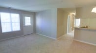 This single-floor residence offers a clean, straightforward layout designed for comfortable everyday living. The main living area greets you with soft, neutral-toned walls painted in a warm gray and beige palette, creating a calm and welcoming atmosphere throughout. Plush wall-to-wall carpeting in a light beige tone adds warmth underfoot and contributes to the overall cohesive aesthetic of the space.

A white-paneled entry door with a window allows natural light to filter into the living area, brightening the room and providing a pleasant connection to the outdoors. The open nature of the main living space ensures a sense of roominess, with clean white baseboards framing the perimeter and providing a crisp contrast to the soft wall colors.

A partial wall divides the living area from an adjoining hallway, which leads further into the home. The hallway connects to additional rooms, suggesting a thoughtful floor plan that separates living and private spaces effectively. Ceiling-mounted lighting fixtures provide consistent illumination throughout.

Overall, the space presents a well-maintained, move-in-ready interior with a neutral design foundation that offers versatility for a variety of furnishing styles and personal touches.