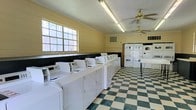 This shared laundry facility is designed to accommodate high-volume use with efficiency and practicality in mind. A long row of white top-load washing machines lines one wall, accompanied by stacked dryer units positioned along the adjacent wall, providing generous capacity for residents. The classic black-and-white checkered vinyl flooring adds a timeless character to the space while remaining easy to maintain. Pale yellow walls with a contrasting dark wainscoting trim give the room a defined, structured appearance. Natural light filters in through a large multi-pane window along one wall, brightening the interior and reducing the reliance on artificial lighting during daytime hours. Two ceiling fans are mounted overhead, promoting air circulation throughout the room and maintaining a comfortable environment during active use. Fluorescent strip lighting ensures the space remains well-lit at all times. A folding table is available for sorting and organizing laundry. The room's straightforward, functional layout maximizes usable floor space while keeping equipment easily accessible. Crown molding along the ceiling adds a subtle architectural detail that elevates the overall appearance of this practical communal space.