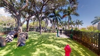 This outdoor dog park offers a well-designed, fully enclosed recreational space tailored specifically for pets. The ground is covered entirely in bright, low-maintenance artificial turf, providing a clean and comfortable surface for dogs to run and play. A secure black iron fence encircles the entire perimeter, ensuring a safe environment where pets can roam freely without concern.

The park is generously shaded by a canopy of mature live oak trees with broad, sprawling branches, complemented by tall, elegant palm trees that contribute a distinctly tropical character. Together, these trees create natural shade across much of the space while allowing ample sunlight to filter through, giving the area an open and airy feel throughout the day.

Several engaging features are distributed throughout the park to encourage activity and exploration. Repurposed oversized tractor tires serve as tunnel obstacles, offering dogs an entertaining crawl-through challenge. A decorative red fire hydrant adds a playful touch, while a green water station is conveniently positioned near the perimeter for easy access. A paved walkway runs along one side of the enclosure, providing a practical path for owners. The combination of thoughtful amenities, natural shade, and vibrant greenery makes this an inviting and functional outdoor space.