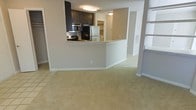 This single-floor residential space offers an open-concept layout that promotes a sense of flow and connectivity between the living and kitchen areas. The kitchen is a standout feature, outfitted with dark espresso cabinetry that provides a striking contrast against the light-colored walls and countertops. Stainless steel appliances, including a refrigerator, microwave, and range, lend a modern, polished look to the space. A peninsula-style breakfast bar serves as both a functional workspace and a natural divider between the kitchen and the living area, making it ideal for casual dining or entertaining. The living area is finished with plush, neutral-toned carpeting, creating a warm and comfortable atmosphere, while the kitchen and adjacent spaces transition to smooth tile flooring. Flush-mount ceiling lighting keeps the space well-lit throughout. Neutral beige and white walls provide a clean, versatile backdrop throughout the home. Additional doorways visible from the main living space suggest connectivity to further rooms, adding to the overall functionality of the floor plan. The cohesive design and open layout make this a practical and inviting living space.