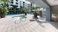 This amenity space is designed to offer a well-rounded recreational experience for its residents. At its centerpiece is a generously sized outdoor swimming pool framed by a clean, tiled pool deck. The pool is complemented by an adjacent bubbling spa, both surrounded by large decorative planters featuring tropical palm trees that lend the area a resort-like atmosphere. Rows of lounge chairs line the deck, providing ample space for sunbathing and relaxation, while the multi-story residential building wraps around the courtyard, creating a sense of enclosure and privacy. The outdoor area is bright and open, bathed in natural light throughout the day.

Connecting to the outdoor space is an indoor recreation and lounge area finished in warm yellow tones with tile flooring throughout. This room is anchored by a full-size ping-pong table and features a large, wrap-around sectional sofa arranged around a patterned area rug and a wooden coffee table. Abstract artwork in cool blue tones adorns the walls, adding a contemporary touch. Recessed lighting illuminates the space evenly. A water fountain and a stocked beverage vending machine provide convenient refreshment options, making this lounge a functional and social hub for residents.