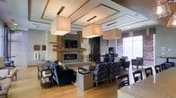This single-floor space exudes a refined, contemporary ambiance designed for both relaxation and social engagement. At the heart of the room stands a striking fireplace framed by a mosaic tile surround and a warm wood mantel, complemented by a built-in media wall with a mounted flat-screen television. The ceiling draws immediate attention with its coffered detailing and a series of large, square fabric pendant lights that cast a warm, even glow throughout the space. Rich hardwood flooring anchors the room and flows seamlessly across the open layout. Multiple seating arrangements — including deep navy sofas, patterned accent chairs, and dark wood dining-style chairs — provide versatile areas for gathering or quiet retreat. Large windows along one wall flood the interior with abundant natural light, softened by horizontal blinds for adjustable ambiance. The open-plan design encourages fluid movement between the lounge and dining areas, creating a sense of spaciousness without sacrificing intimacy. Thoughtful decorative accents, including sculptural objects and greenery, add personality and warmth to this well-appointed, functional space.